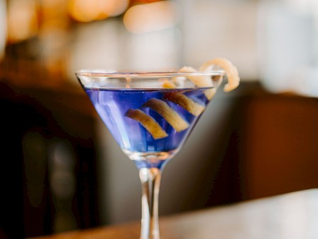 A martini glass with a blue cocktail, garnished with a lemon twist, on a coaster in a warmly lit setting.
