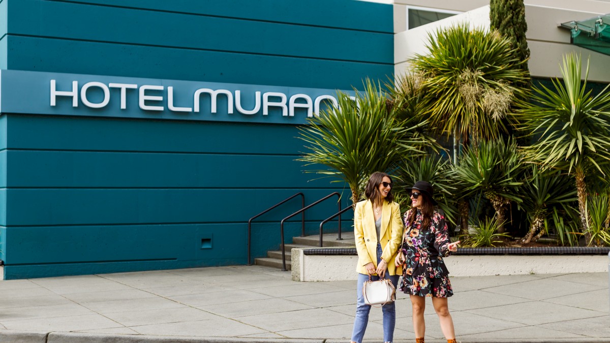 Two stylish people stand outside a blue Hotel Murano, posing in front of palm plants and steps by the entrance.