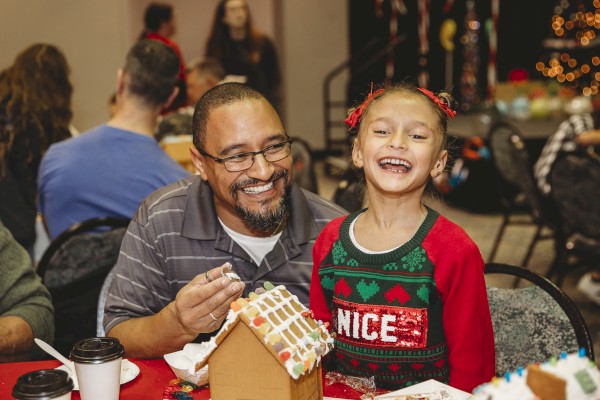 A smiling man helps a girl decorate a gingerbread house at a festive table, cups and cookies around, warm holiday glow, joyful moment.