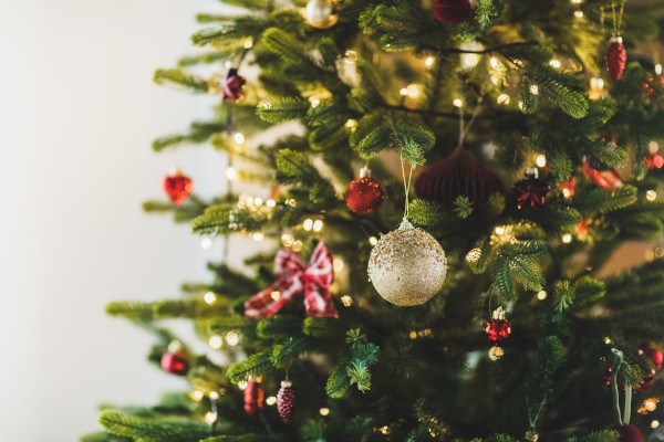 A Christmas tree decorated with lights, red ornaments, ribbons, and a silver bauble hanging in the foreground.