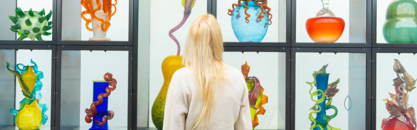 A woman observes colorful glass sculptures displayed in a grid-like window at an art museum.