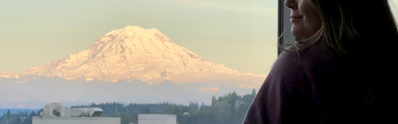 A woman watches Mount Rainier from a window, with the snow-capped mountain visible in the background during sunset or sunrise.