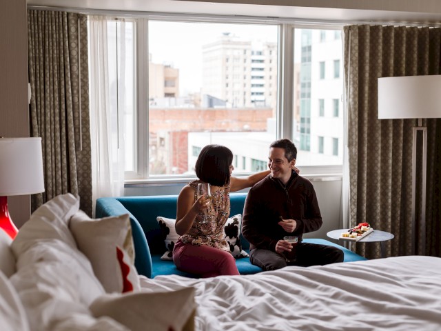 A woman and man sit on a blue sofa, sharing drinks and smiling in a bright hotel room with city view through large windows.
