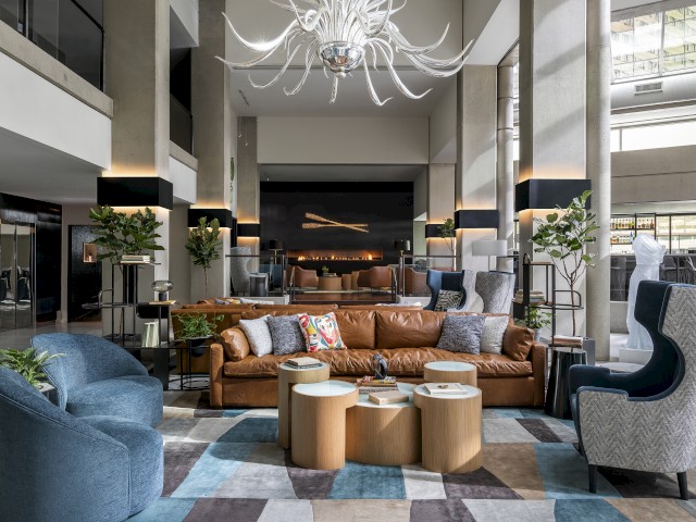 A stylish hotel lobby with modern furniture, sofas, armchairs, plants, and a large decorative chandelier in a spacious setting.