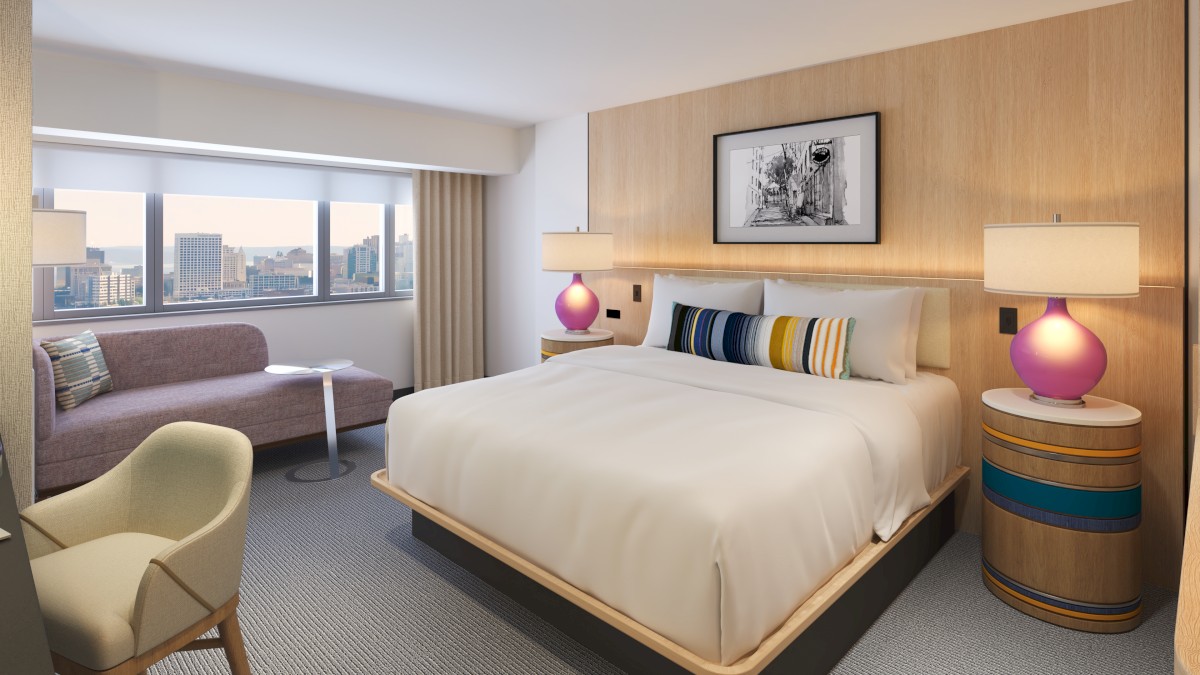 A modern hotel room with a large bed, side tables, colorful lamps, a sitting area, and city views through large windows.