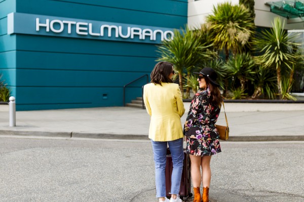 Two women with luggage smiling and talking outside Hotel Murano, with tropical plants and a blue building in the background.