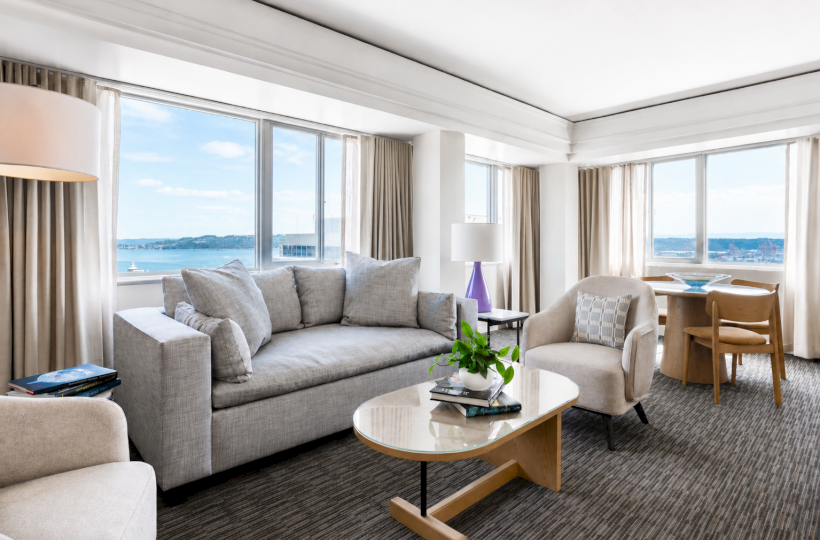 A bright, modern living room with large windows, beige furniture, a coffee table, and a view of water outside.