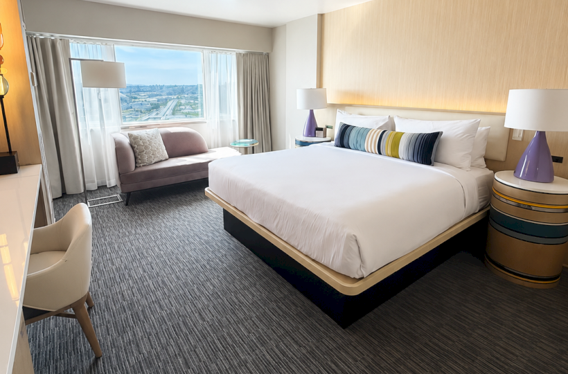 This hotel room has a bed, side lamps, a window with city view, a sofa, and a desk, featuring neutral tones and modern decor.