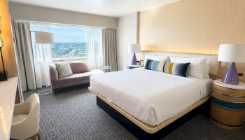 A modern hotel room features a large bed, a bench by the window, side tables with lamps, and a city view outside.