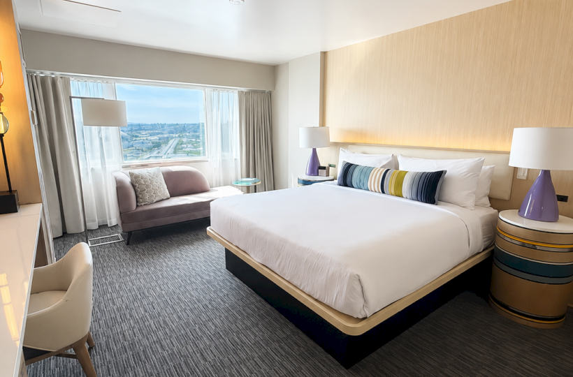 A modern hotel room features a large bed, a bench by the window, side tables with lamps, and a city view outside.
