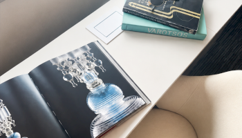 A desk with books, sunglasses, a lamp, and an open art book featuring glassware, next to a beige chair at a window.