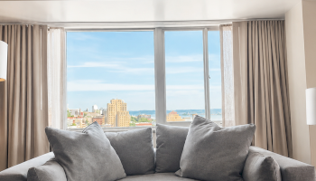 A stylish living room with a grey sofa, a round coffee table, a green plant, bookshelf, and city skyline view through large windows.