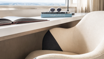A cozy workspace by the window with a beige and black chair, a white desk, reading glasses, and a lamp with a view of the sky.