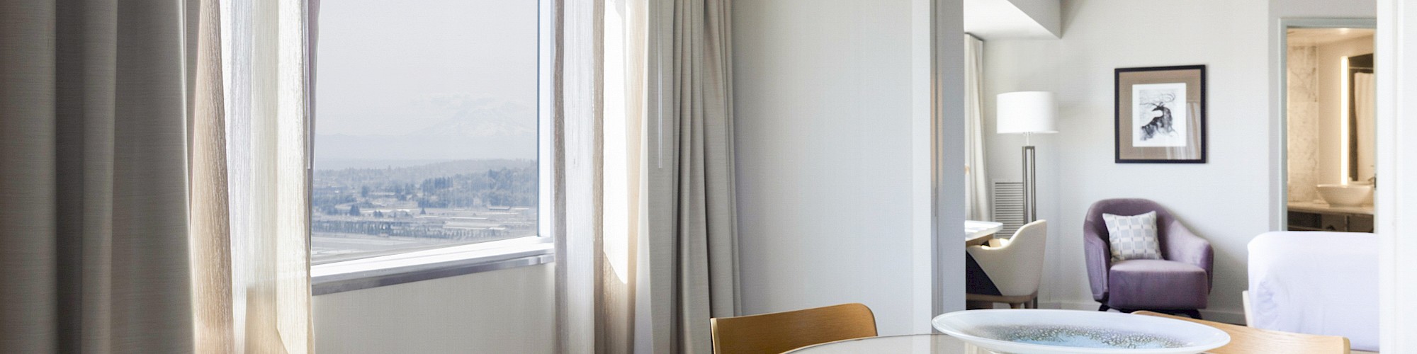 A stylish hotel suite with a round wooden dining table, chairs, a large window view, and a cozy living area with artwork and lighting.