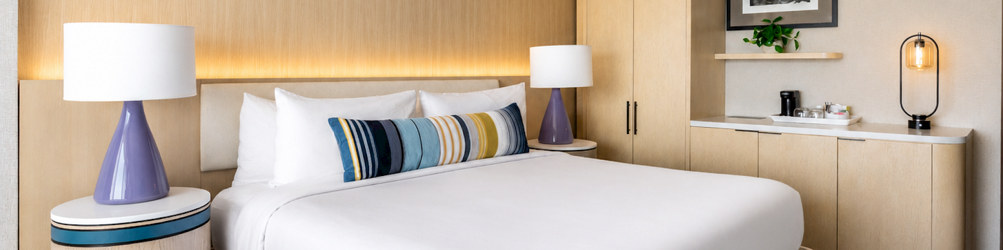 A modern hotel room with a large bed, bedside lamps, decorative pillow, and a minimalist wooden cabinet with a small coffee/tea station.