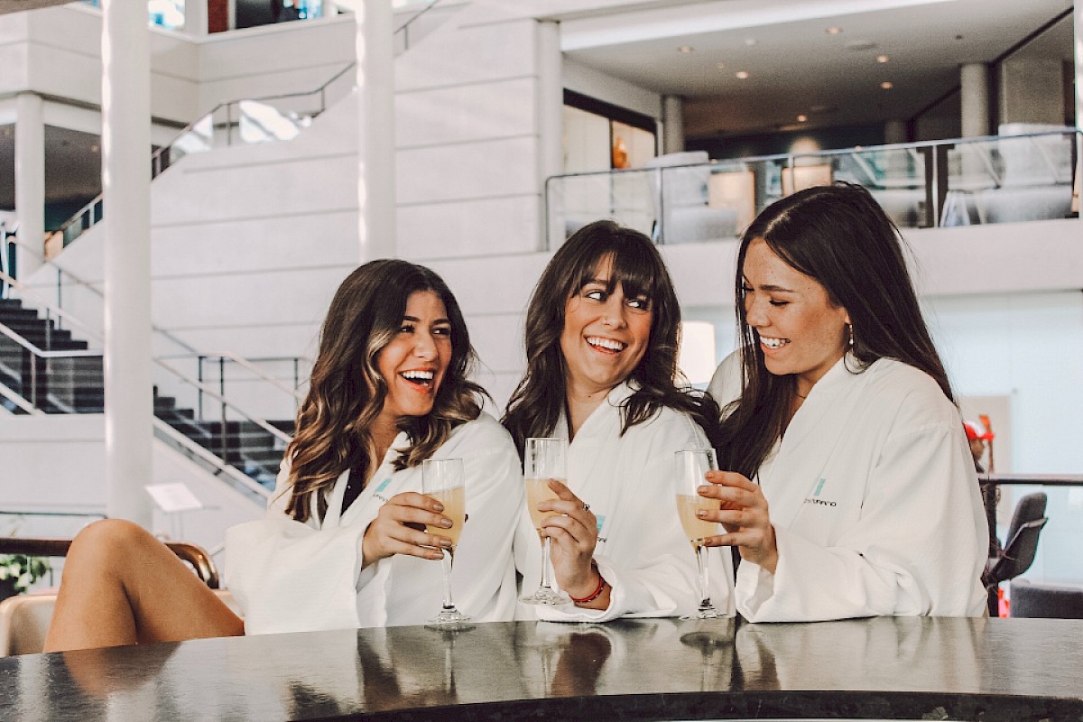 Three women in white robes are enjoying drinks and laughing together at a modern indoor location, likely celebrating or relaxing.