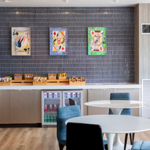 This image shows a cozy refreshment area with snack shelves, a mini fridge, tables, chairs, and abstract artwork on the wall.