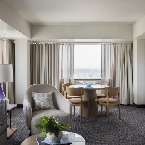 A bright, modern hotel room with a sitting area, round dining table, chairs, a sofa, a lamp, a potted plant, and a window with curtains.