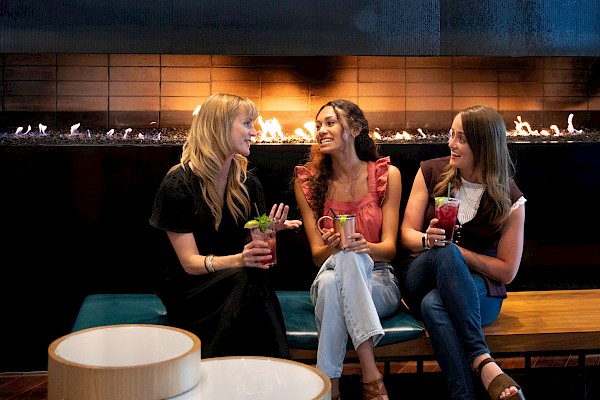 Three women are sitting on a bench, chatting and holding colorful drinks in a modern, cozy lounge with a fireplace in the background.