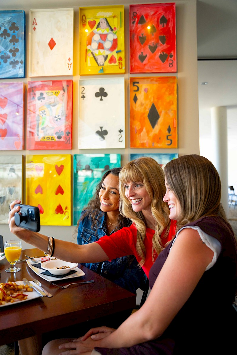 Three women are taking a selfie at a dining table with food and drinks, in front of colorful artwork featuring playing cards.