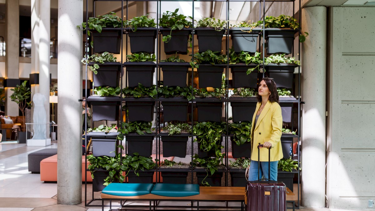 A woman with dark hair, yellow coat, and suitcase waits with luggage in front of a green plant wall inside a modern building.
