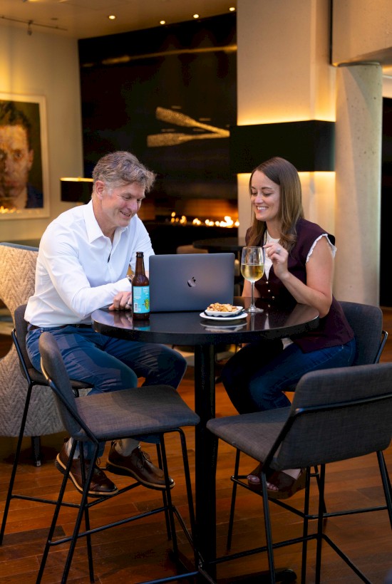 A man and woman sit at a black table, sharing drinks and looking at a laptop in a cozy, modern lounge with warm lighting and artwork.