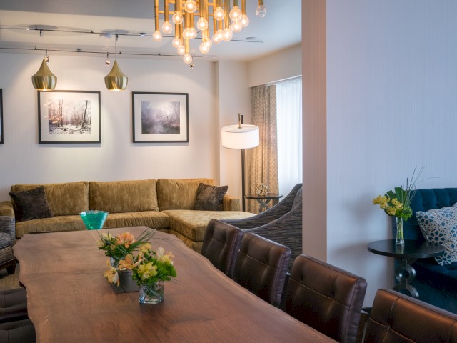 A cozy, stylish living room with a wooden dining table, plush sofas, framed art, and elegant lighting fixtures.