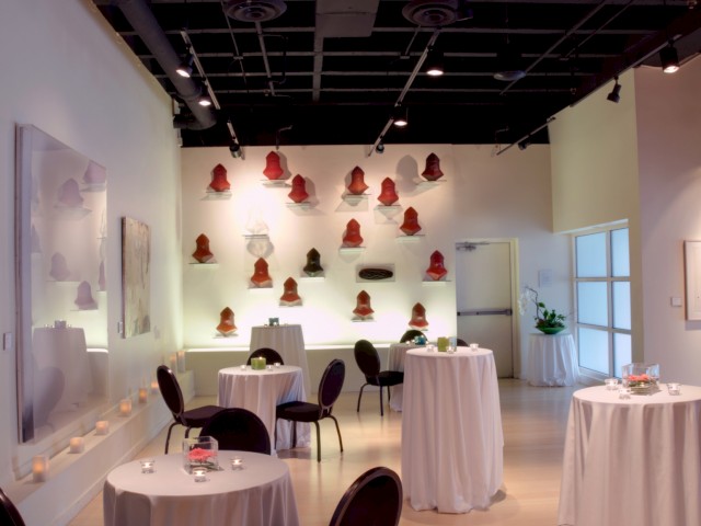 This is an elegant, softly lit event space with round tables covered in white cloths, candles, and a wall display of red and black sculptures.