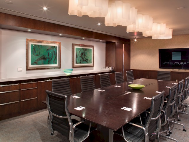 This is an elegant conference room with a large wooden table, black chairs, wall art, and a TV displaying 'Hotel Murano'.