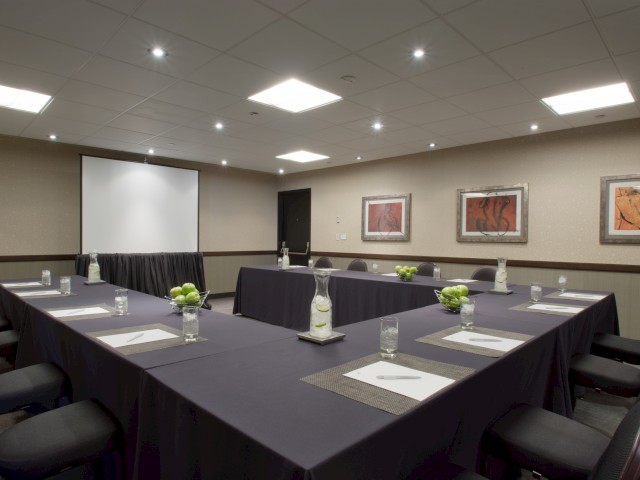 A conference room with a U-shaped arrangement of black tables, chairs, artwork on the walls, and a projection screen.